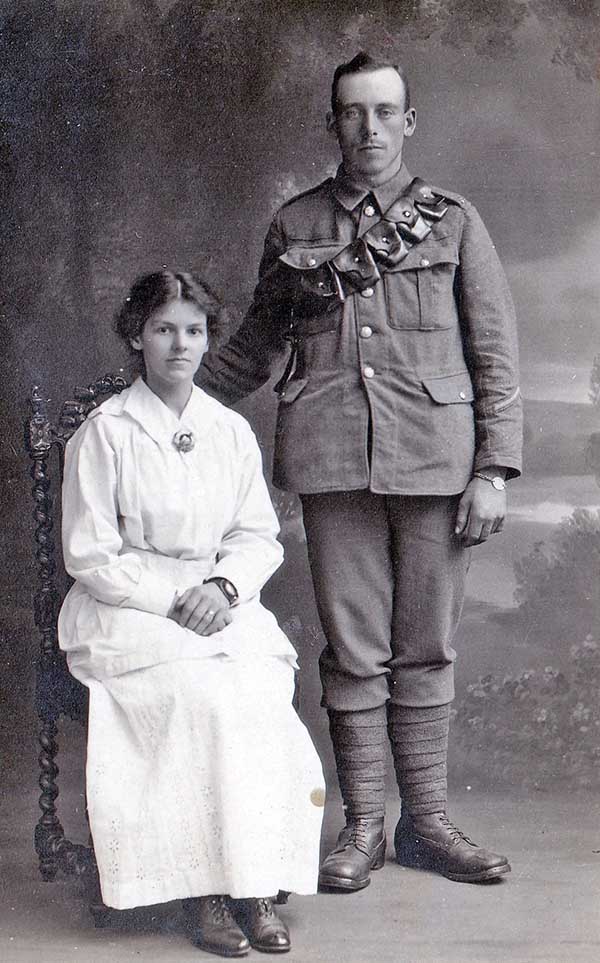 black and white photograph of woman in a long light-coloured skirt and top sitting on a chair, with a man in Army uniform standing next to her. The backdrop suggests the picture was taken in a photographer's studio.