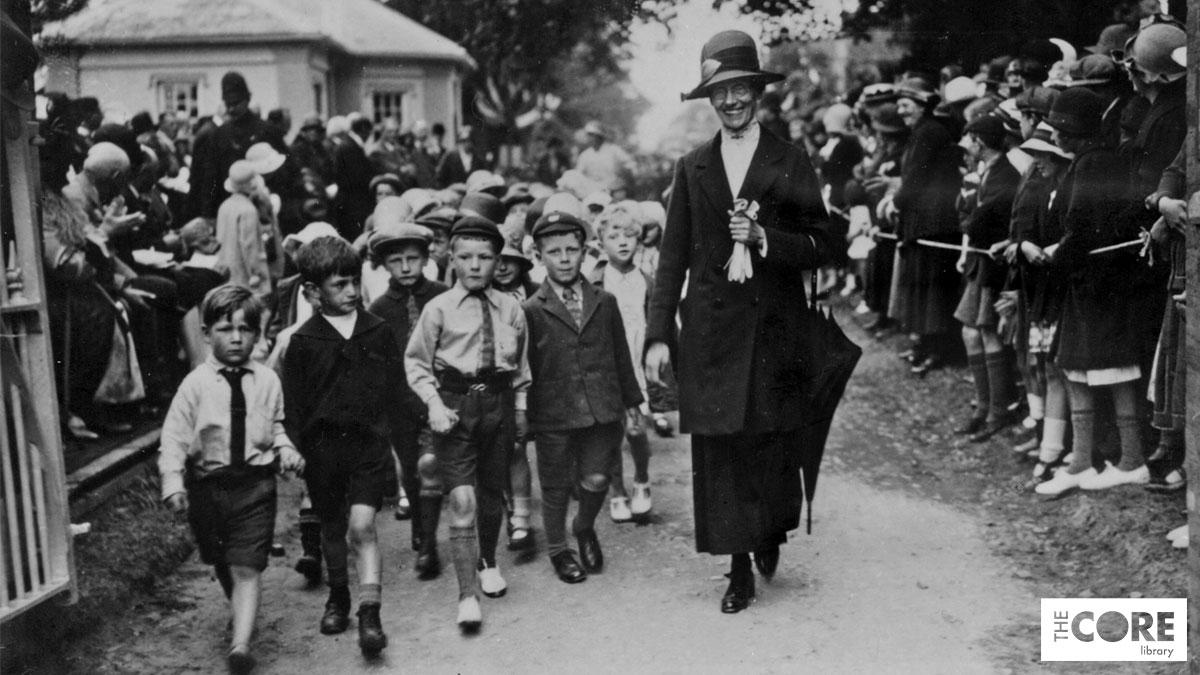 A black and white photo from 1926 showing a group of young children with their teacher walking along a path lined with spectators.